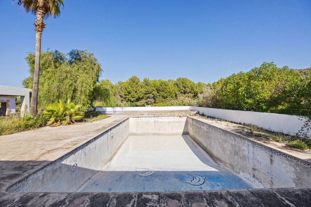 Piscina rectangular de grandes dimensiones para rehabilitar, rodeada de una amplia terraza de piedra y vegetación mediterránea bajo el cielo despejado.