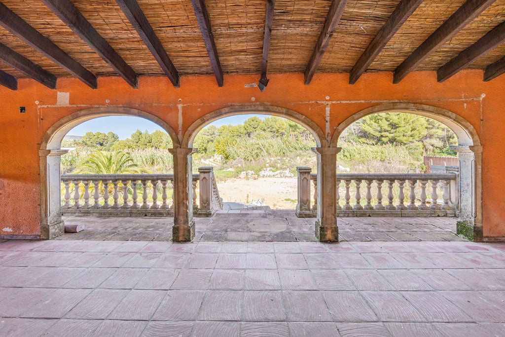 Terraza cubierta con arcos de piedra tosca, techo de vigas de madera y cañizo, con vistas despejadas al entorno natural mediterráneo y balaustrada clásica.