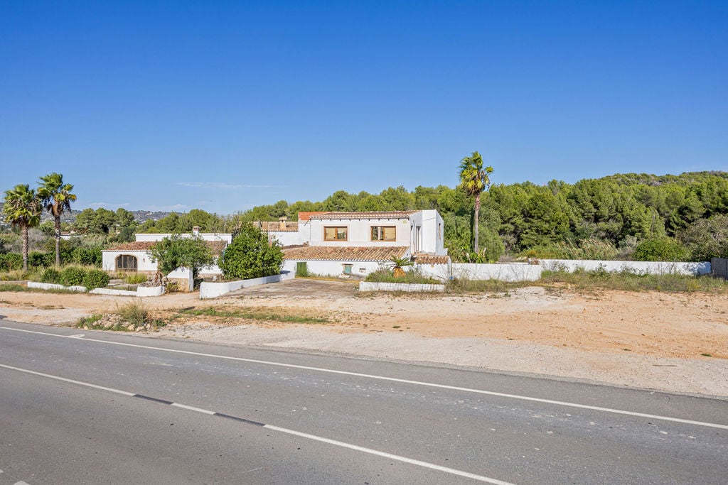 Vista exterior de la finca tradicional con fachada blanca y tejados de teja cerámica, situada junto a una carretera de acceso y rodeada de vegetación mediterránea.
