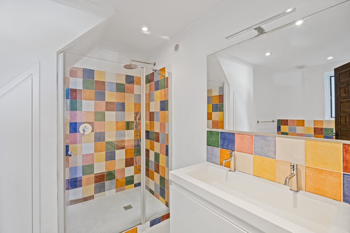 Baño moderno con lavabo doble integrado y ducha con mampara de cristal, destacando el revestimiento de azulejos cerámicos multicolores e iluminación empotrada.