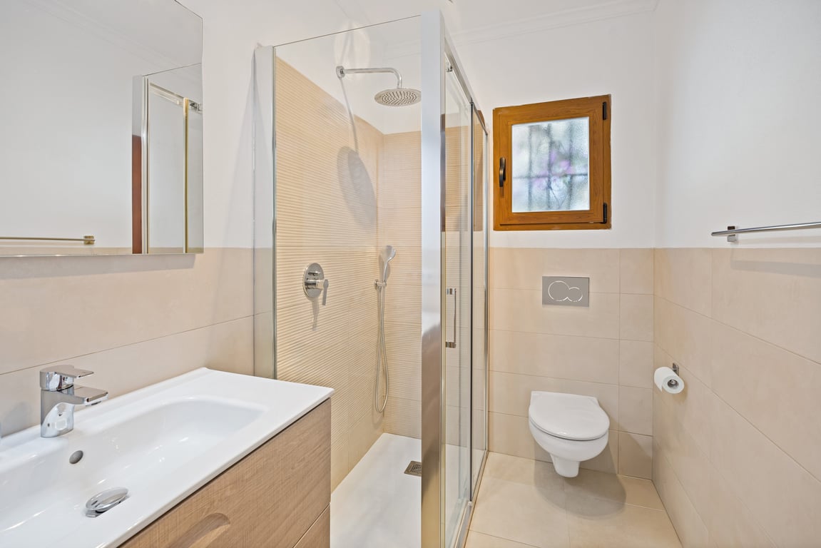 Baño moderno reformado con ducha de obra, mampara de cristal, lavabo con mueble de madera clara y sanitarios en tonos crema.