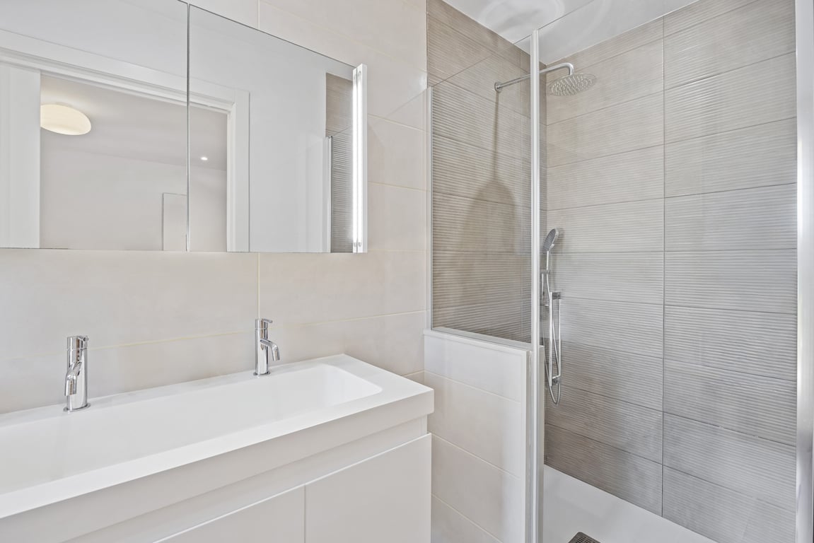 Baño moderno reformado con lavabo de doble grifería, mueble blanco minimalista y ducha con mampara de cristal y azulejos texturizados en tonos neutros.