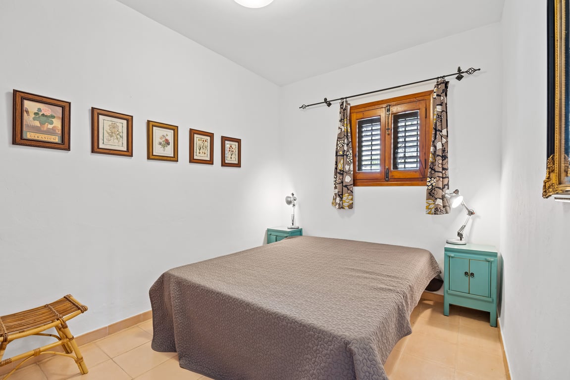 Dormitorio con cama doble, mesitas de noche en color turquesa y ventana de madera con persianas tradicionales. Paredes blancas decoradas con cuadros botánicos.
