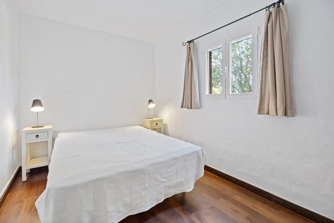 Dormitorio luminoso con suelos de madera, paredes blancas y ventana de doble acristalamiento con vistas al exterior. Incluye mesitas de noche y lámparas funcionales.