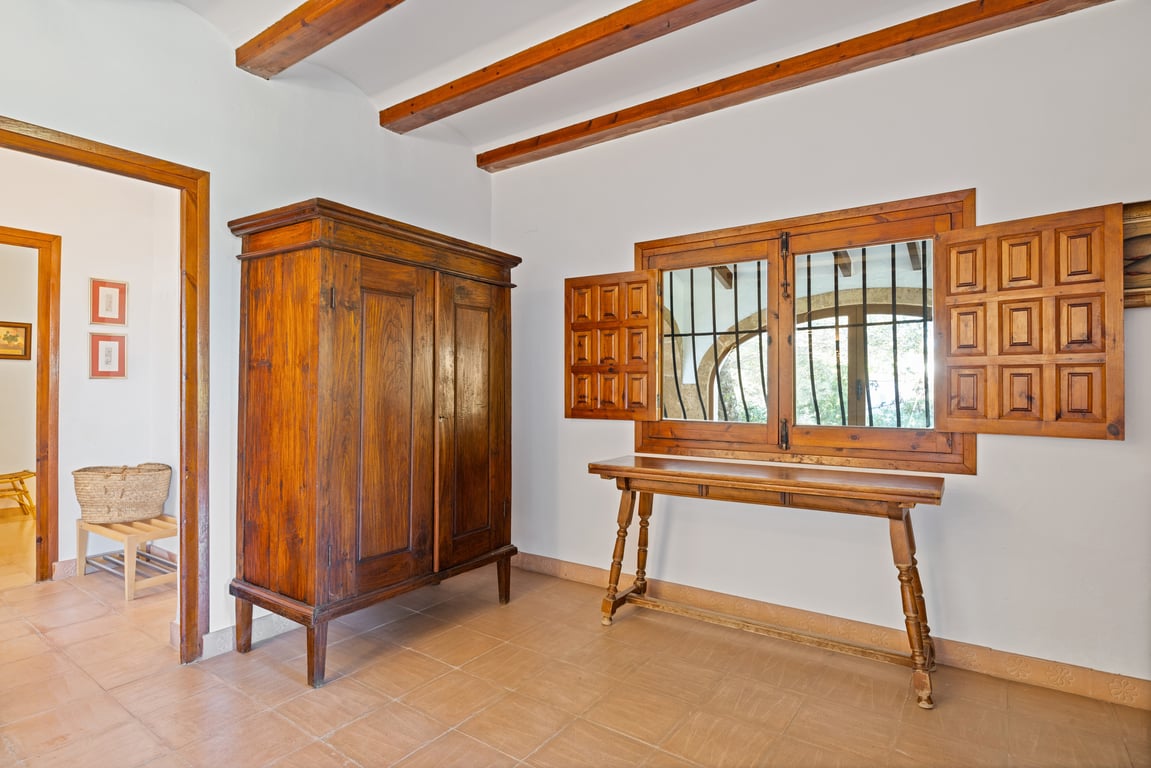 Entrada con vigas de madera vista, suelo de baldosa cerámica artesana y mobiliario tradicional. La ventana incluye contraventanas clásicas y rejas de seguridad.