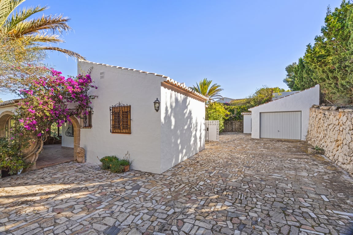 Entrada exterior con suelo de piedra irregular, garaje independiente y fachada blanca decorada con buganvilla rosa y arcos de piedra Tosca tradicional.