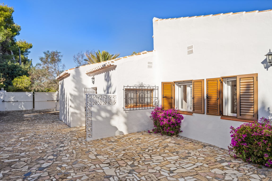 Exterior de la villa con fachada blanca, contraventanas de madera, rejas de forja y suelo de piedra natural irregular junto a buganvillas.