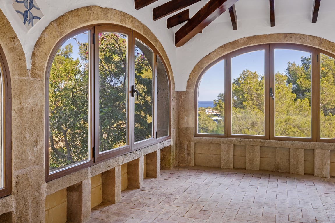 Naya acristalada con arcos de piedra Tosca, vigas de madera a la vista y suelo de baldosa artesanal. Los ventanales ofrecen vistas al mar y al entorno natural de Jávea.