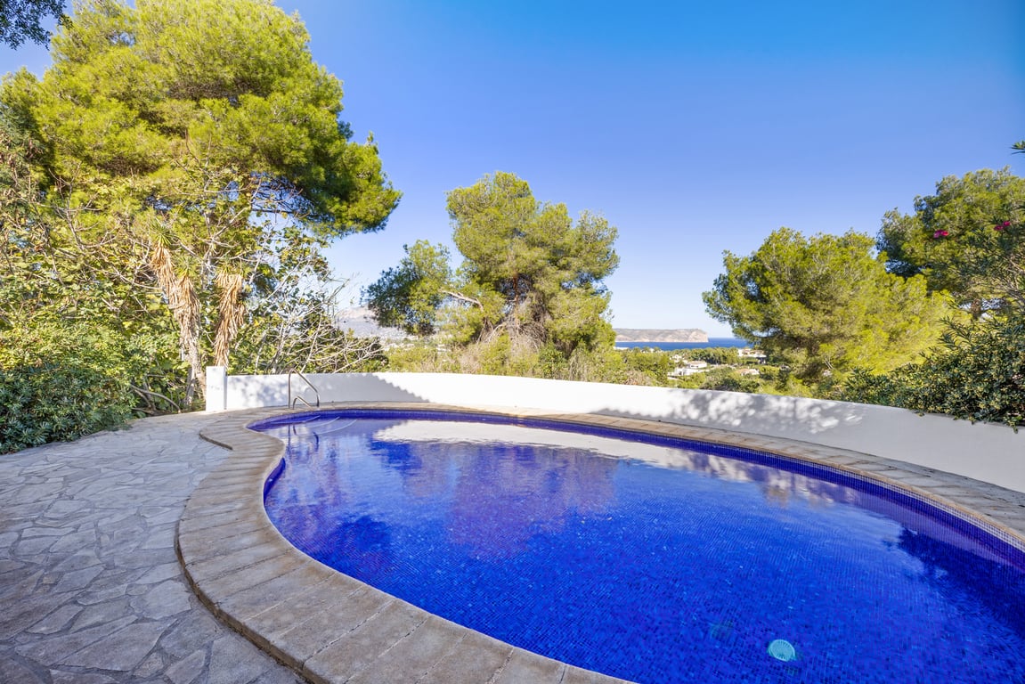 Piscina privada de forma curva con gresite azul, rodeada de una terraza de piedra natural y pinos maduros con vistas lejanas al mar Mediterráneo.