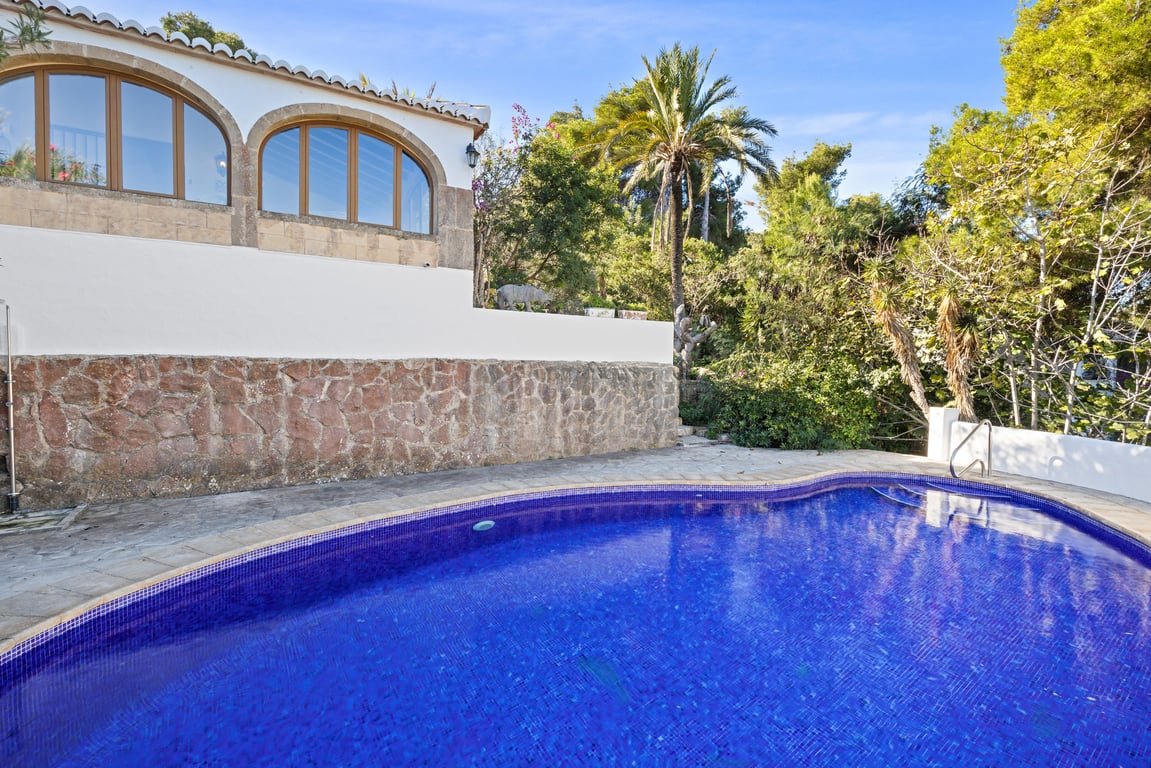 Piscina privada rodeada de una terraza de piedra y muros de contención rústicos, con la naya de arcos acristalados y vegetación mediterránea al fondo.