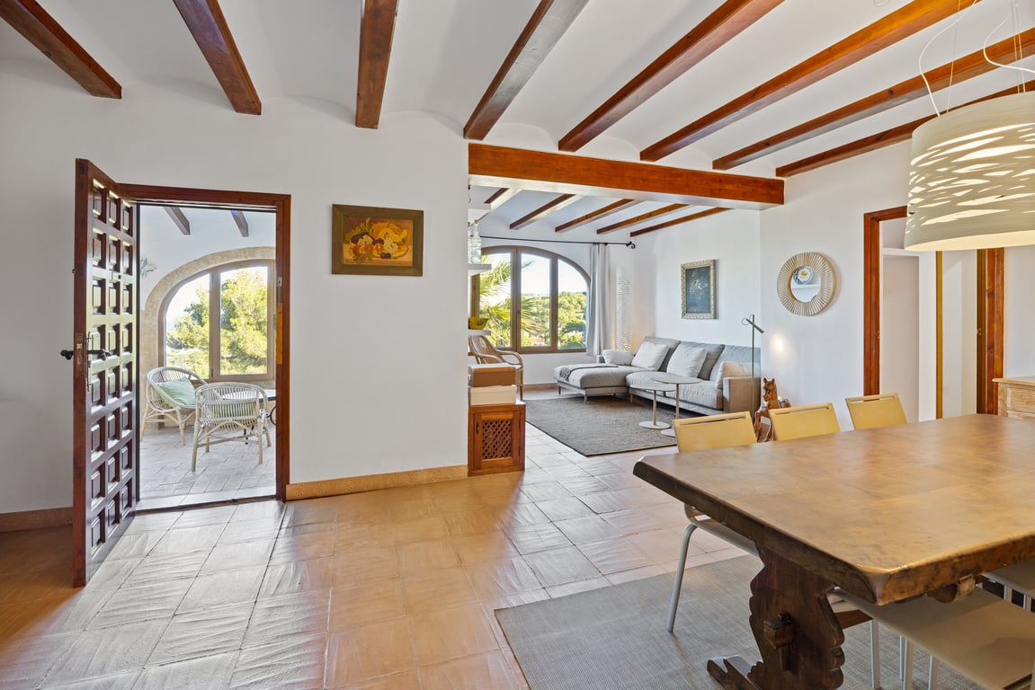 Salón-comedor tradicional con vigas de madera, suelo de barro artesanal y acceso a una naya cubierta con vistas exteriores y arcos de piedra Tosca.