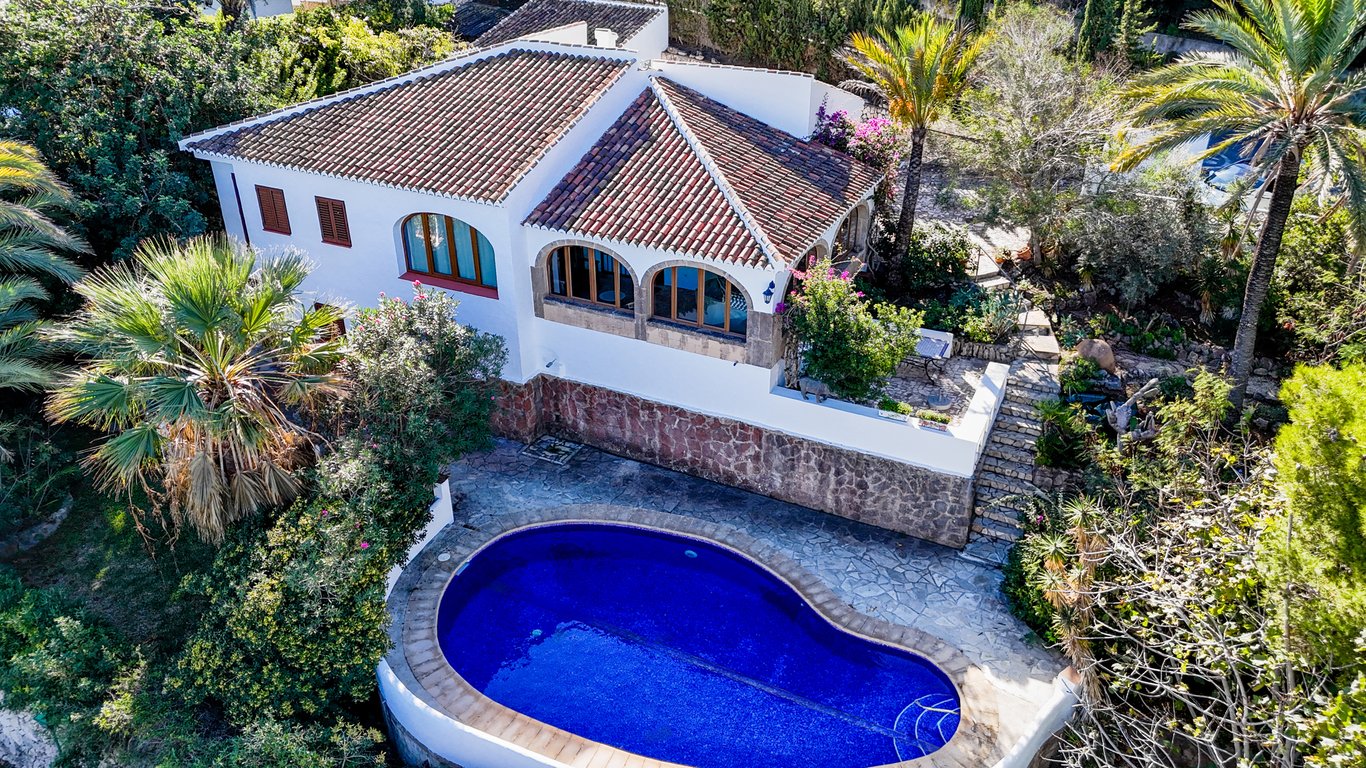 Vista aérea de la villa con piscina privada en forma de riñón, fachada blanca con base de piedra y naya acristalada con arcos de piedra Tosca rodeada de vegetación.