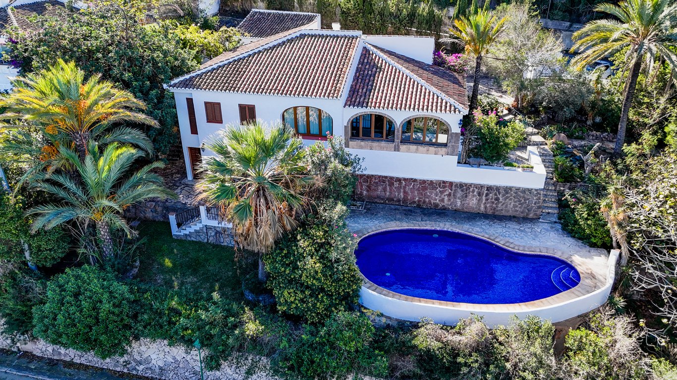 Vista aérea de la villa con su piscina privada de forma riñonada, rodeada de vegetación mediterránea, palmeras y una naya con arcos de piedra Tosca tradicional.