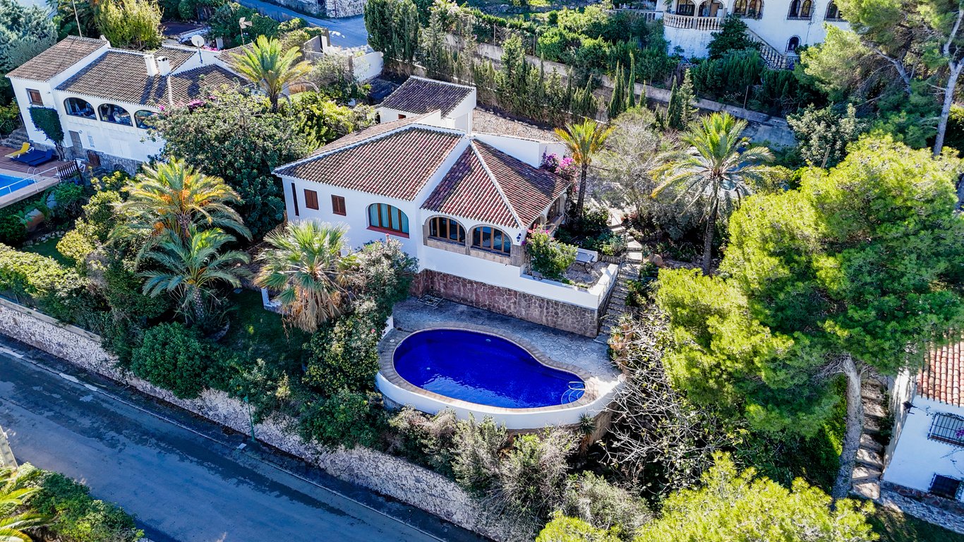 Vista aérea de la villa tradicional con piscina en forma de riñón, rodeada de vegetación mediterránea, palmeras y muros de piedra natural en una zona residencial.