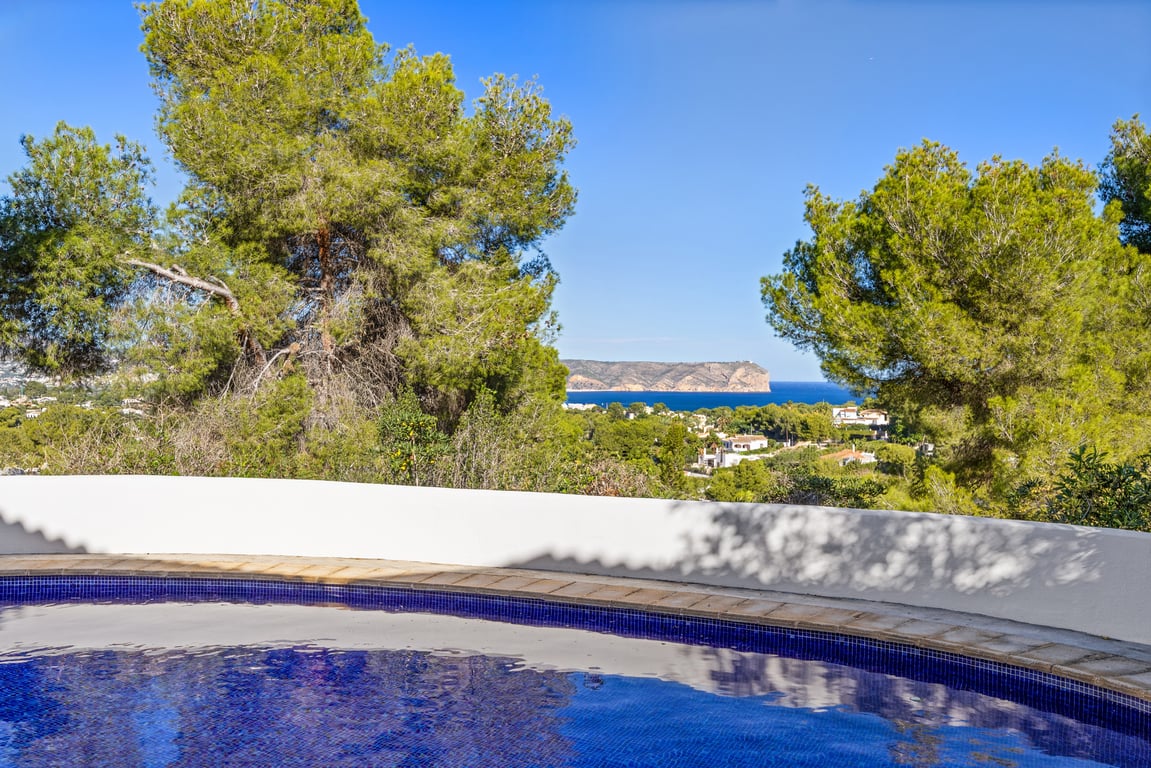 Vista desde la piscina privada hacia el mar Mediterráneo y el cabo, rodeada de pinos maduros y un muro curvo blanco tradicional.