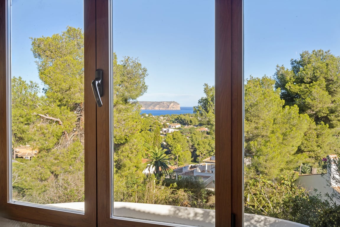 Vista panorámica al mar y al cabo desde una ventana con carpintería de efecto madera y doble acristalamiento, rodeada de pinos mediterráneos.