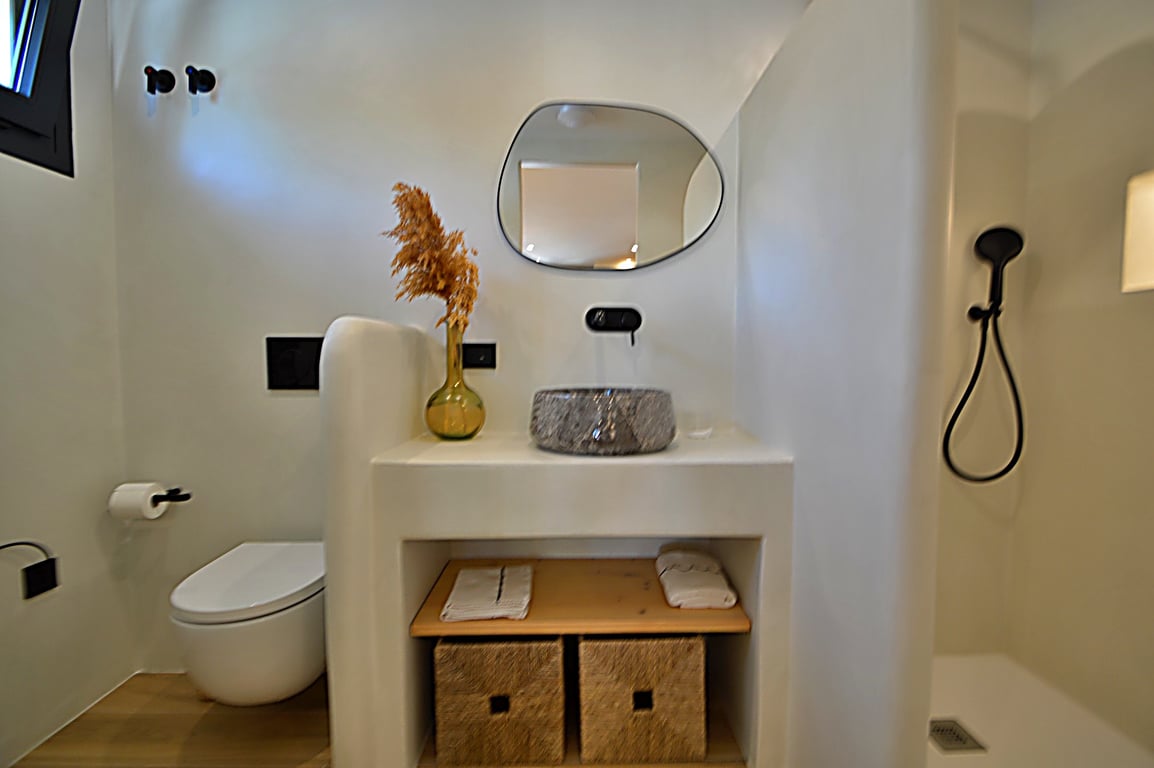 Baño de estilo ibicenco con paredes de microcemento, lavabo de piedra sobre encimera de obra, grifería negra mate y espejo orgánico. Incluye ducha de obra y estantería de madera.