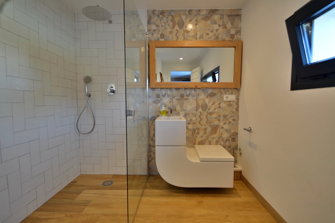 Baño moderno con ducha de obra, mampara de cristal y un lavabo integrado con inodoro de diseño. Destaca la pared de azulejos geométricos y el espejo con marco de madera natural.