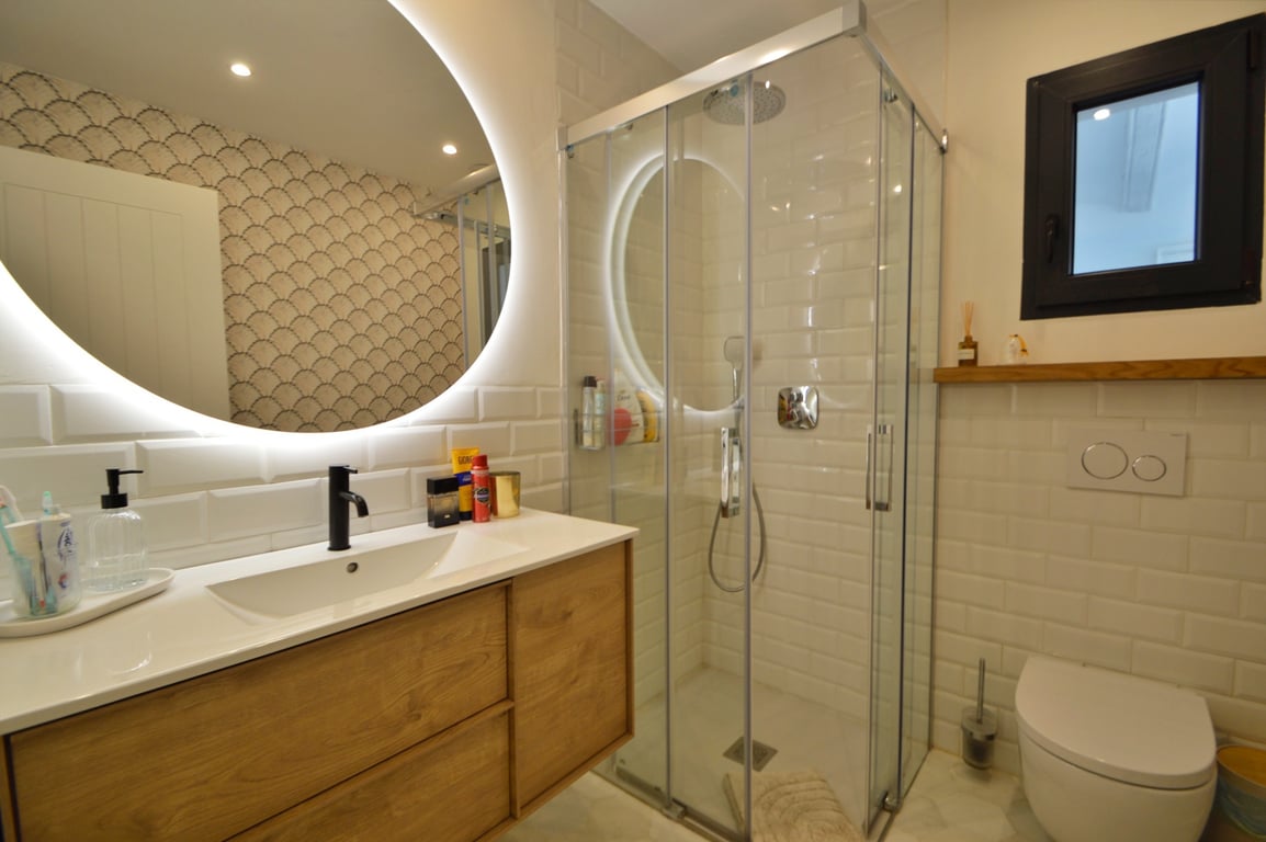 Baño moderno con espejo circular retroiluminado, mueble de madera con grifería negra y ducha de esquina con azulejos tipo metro.