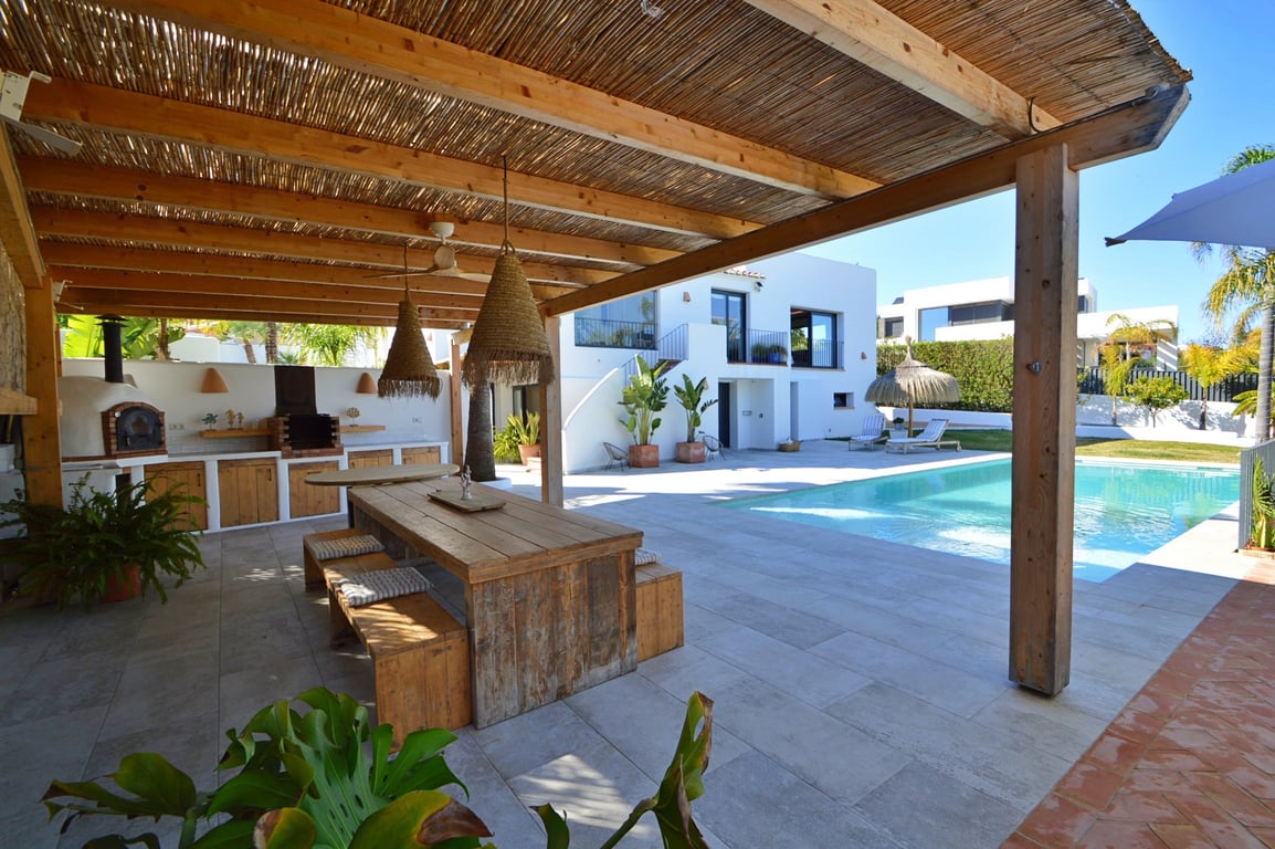 Cocina exterior bajo pérgola de madera con horno de leña, comedor rústico y vistas a la piscina de agua salada y la fachada blanca de estilo ibicenco.