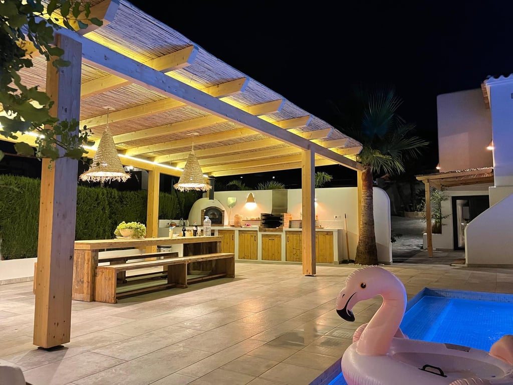Cocina exterior iluminada con horno de leña, barbacoa y zona de comedor bajo una pérgola de madera y cañizo junto a la piscina.