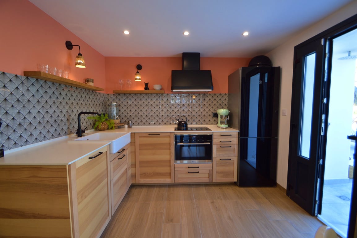 Cocina moderna con armarios de madera clara, fregadero de estilo rústico y salpicadero geométrico. Combina electrodomésticos negros con una pared terracota y estantes flotantes.
