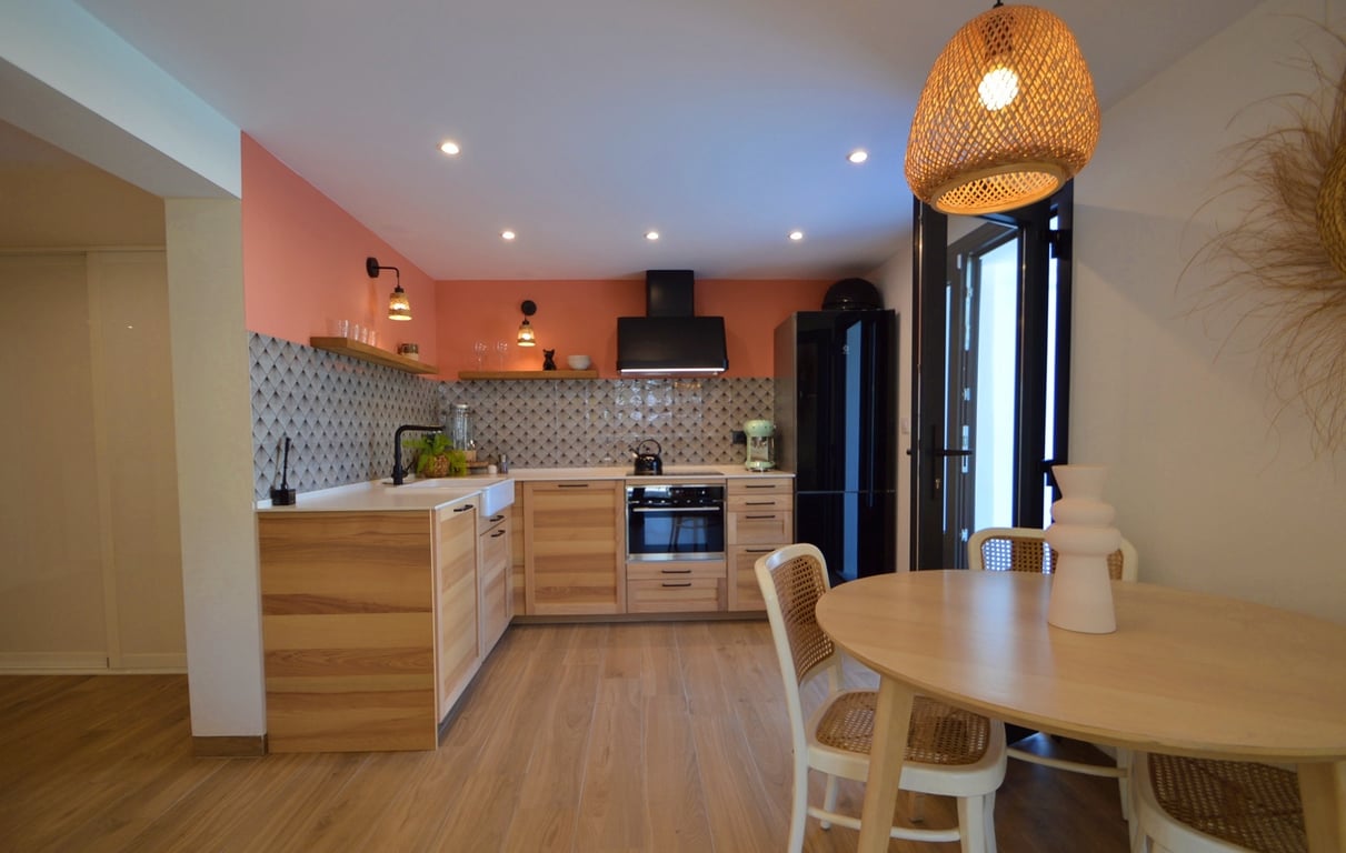 Cocina moderna con armarios de madera, pared de acento terracota y zona de comedor integrada con mesa redonda, sillas de rejilla y una lámpara colgante de fibras naturales.