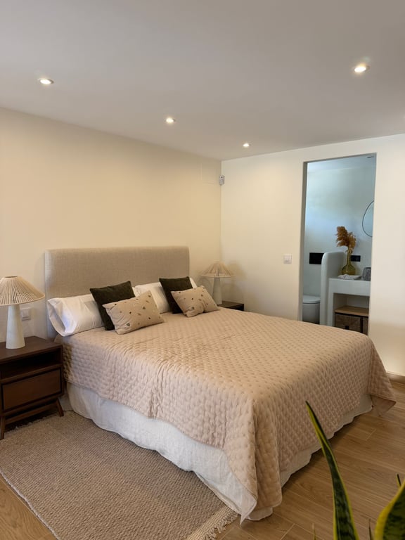 Dormitorio luminoso con cama doble, cabecero tapizado y mesitas de madera. Incluye baño en suite integrado con acabados modernos y suelo de gres efecto madera.