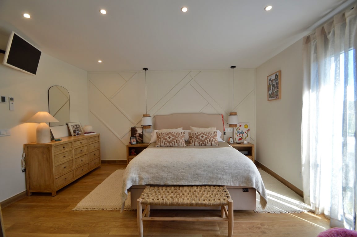 Dormitorio principal con pared de diseño geométrico, suelos de madera y mobiliario en tonos naturales. Incluye iluminación empotrada, lámparas colgantes y abundante luz natural.