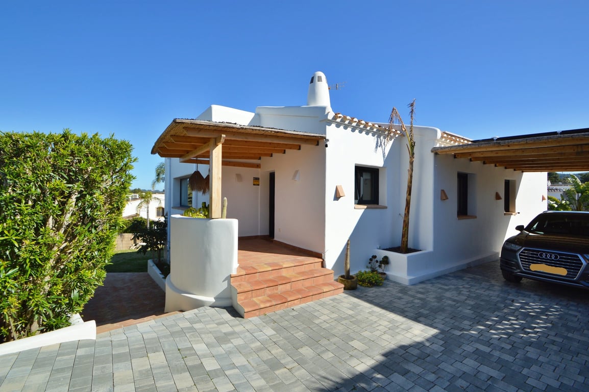 Entrada principal de estilo ibicenco con porche de madera y cañizo, acceso mediante escalones cerámicos y zona de aparcamiento pavimentada con carport lateral.
