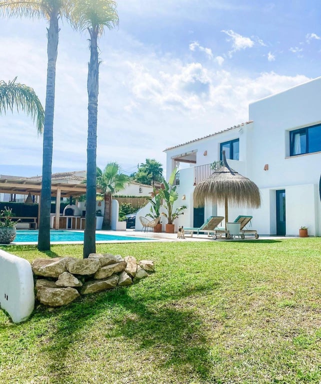Exterior de estilo ibicenco con jardín de césped, piscina y zona de relax bajo sombrilla de brezo. Al fondo destaca la cocina exterior con horno de leña.