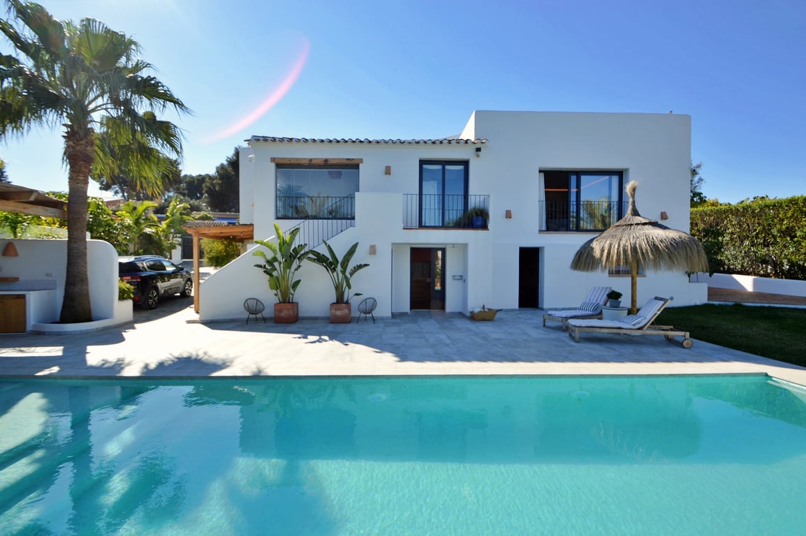 Fachada blanca de estilo ibicenco frente a una amplia piscina de agua salada, con terraza de piedra, palmeras y zona de descanso con sombrilla de brezo.