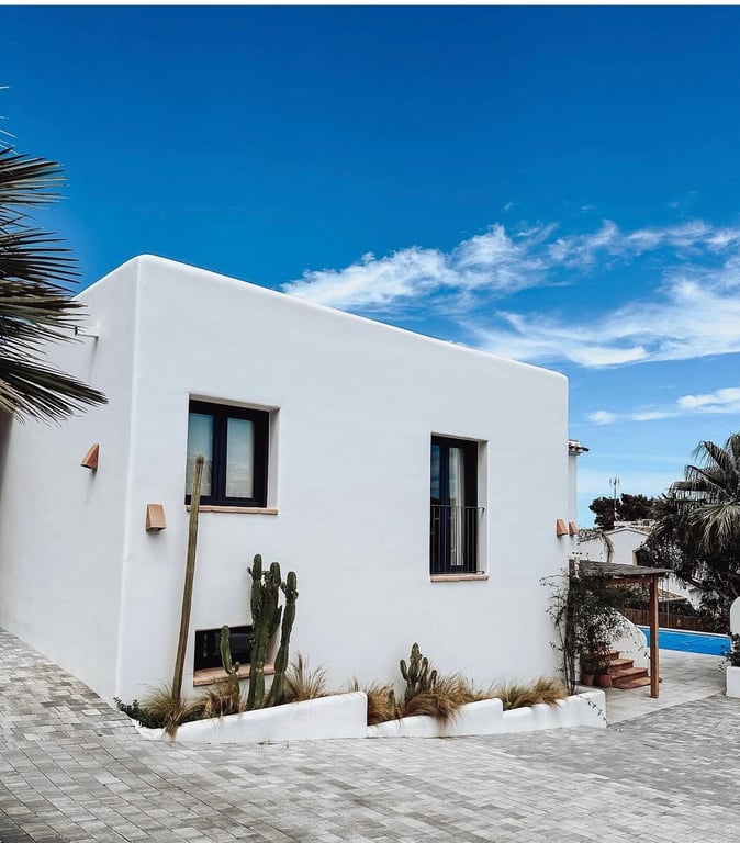 Fachada de estilo ibicenco con paredes blancas lisas, ventanas de carpintería oscura y un jardín de cactus integrado junto al acceso pavimentado con piedra.