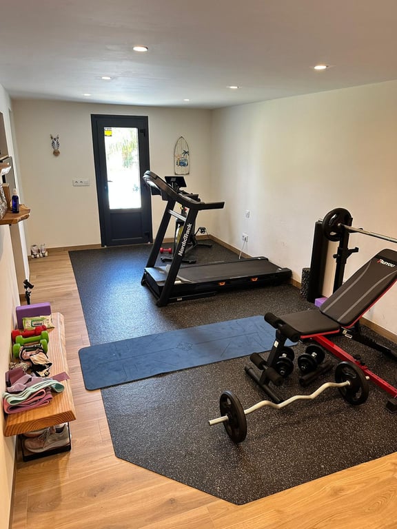 Gimnasio privado equipado con cinta de correr, pesas y zona de yoga, con suelo de madera, iluminación empotrada y acceso directo al exterior.