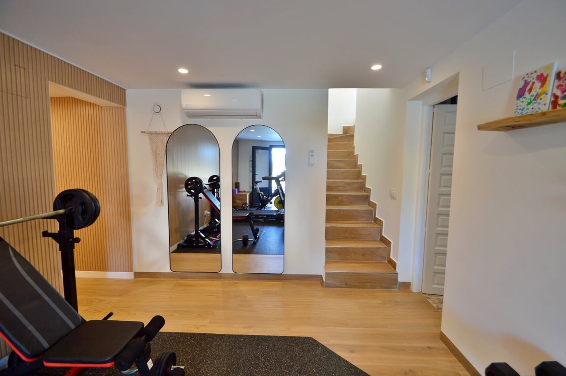 Gimnasio privado equipado con espejos de arco, suelos de madera, aire acondicionado y una escalera de acceso, combinando funcionalidad y diseño moderno.