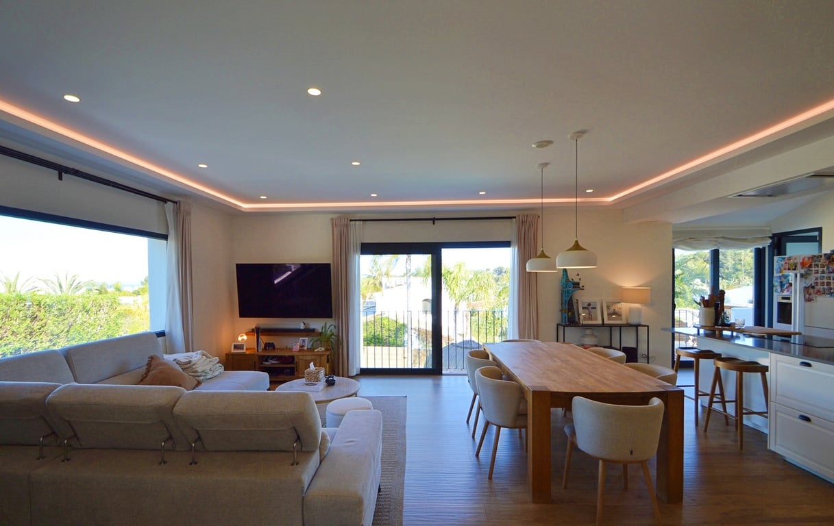 Salón-comedor de concepto abierto con iluminación LED perimetral, suelos de madera y amplios ventanales que integran el paisaje exterior con el interior moderno.