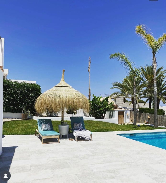 Terraza de piedra clara junto a la piscina de agua salada, equipada con tumbonas turquesa y sombrilla de brezo en un entorno ajardinado con palmeras.