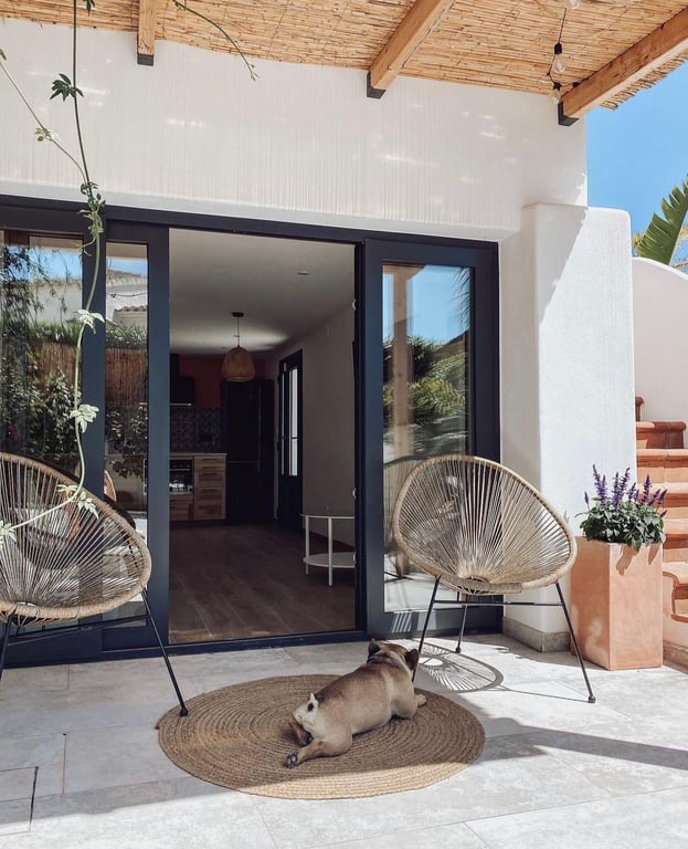 Terraza sombreada con techo de cañizo y vigas de madera, conectada al interior mediante ventanales de carpintería oscura y suelo de piedra natural.