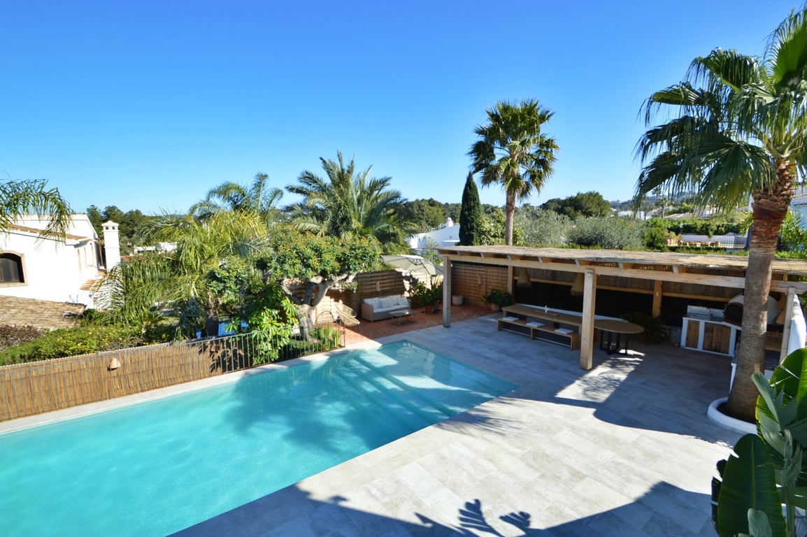 Vista elevada de la piscina de agua salada y terraza de piedra, con una pérgola de madera que alberga el comedor exterior y la cocina con horno de pizza.