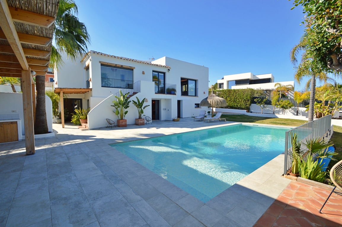 Vista exterior de la villa con piscina rectangular rodeada de una amplia terraza de piedra clara, pérgola de madera y fachada blanca de estilo ibicenco con naya acristalada.