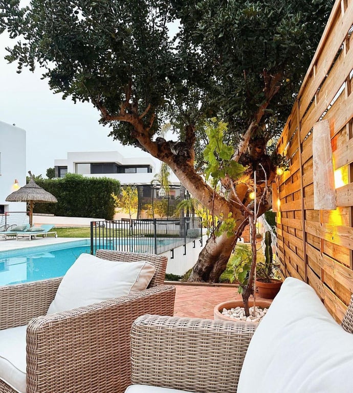 Zona de estar exterior con mobiliario de mimbre y cojines blancos, situada sobre suelo de terracota junto a una valla de madera con iluminación ambiental y vistas a la piscina.