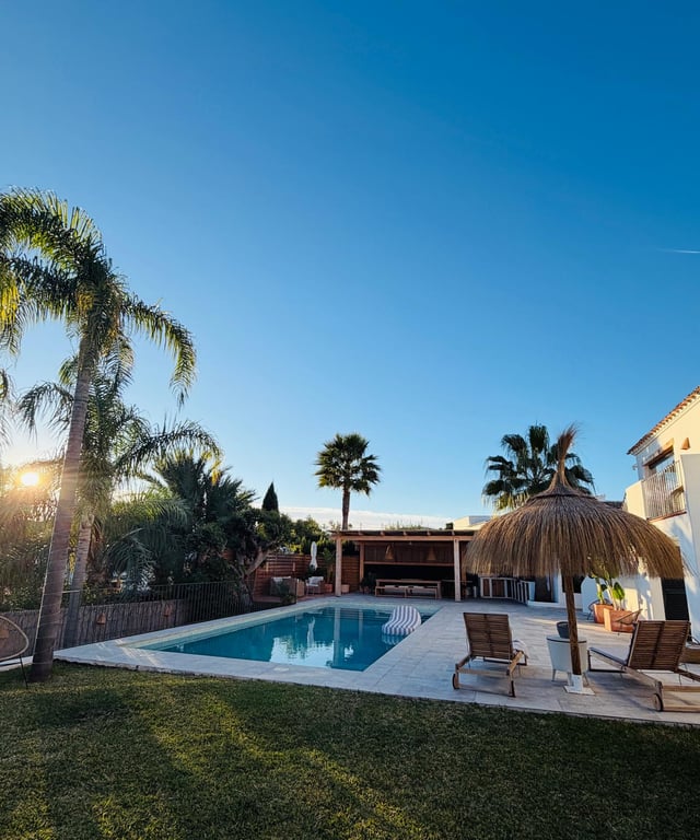 Zona exterior con piscina rodeada de terraza de piedra, sombrilla de brezo y jardín mediterráneo. Al fondo, un porche cubierto con cocina exterior y comedor bajo las palmeras.