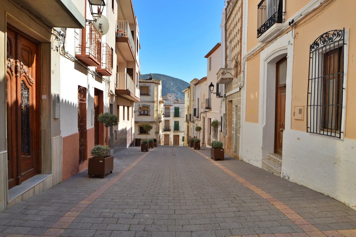 Calle peatonal adoquinada en el centro del pueblo, flanqueada por fachadas tradicionales con balcones de forja y vistas directas a la montaña al fondo.