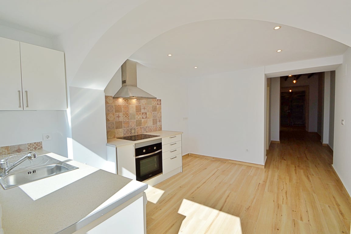 Cocina renovada con armarios blancos, salpicadero de azulejos decorativos y electrodomésticos integrados. Destaca el suelo de madera clara y el arco que conecta con el salón.