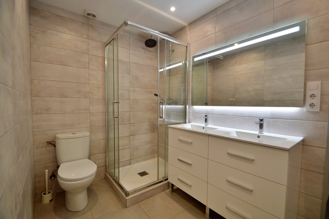 Cuarto de baño moderno con lavabo doble, mueble blanco de seis cajones, espejo retroiluminado y cabina de ducha acristalada con azulejos en tonos crema.