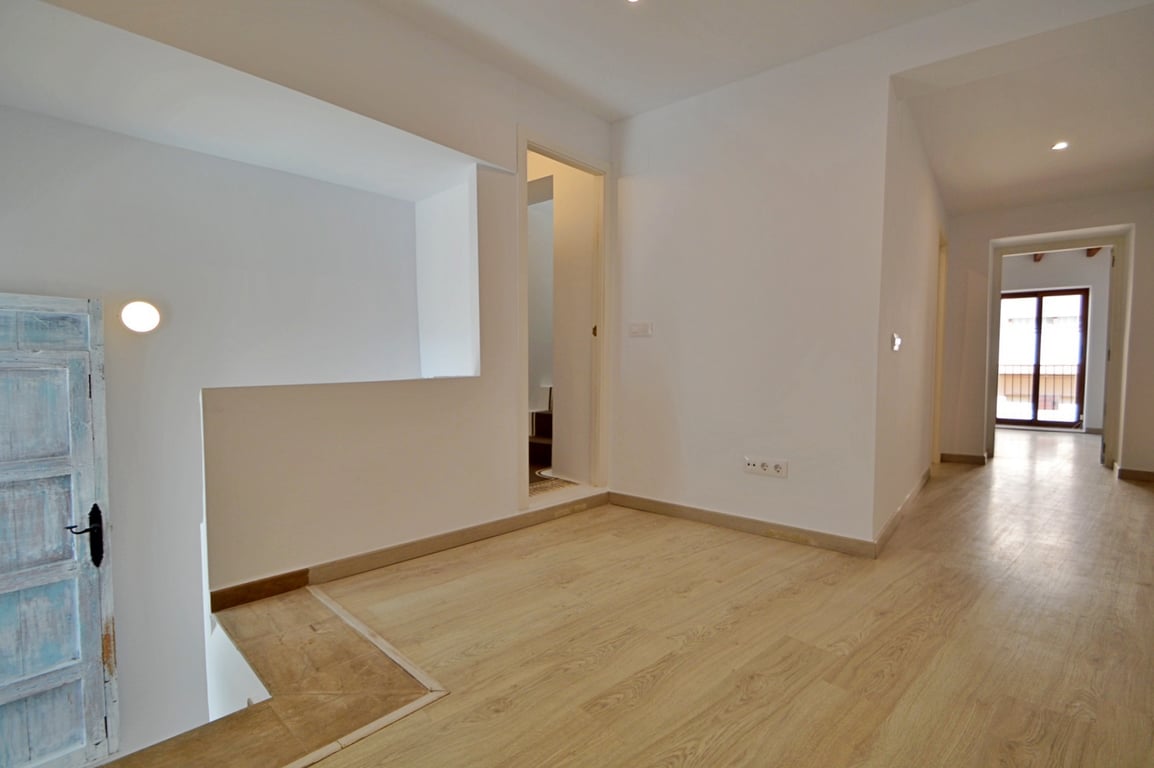 Distribuidor luminoso con suelos de tarima clara, paredes blancas y una puerta rústica restaurada que aporta carácter al espacio renovado.