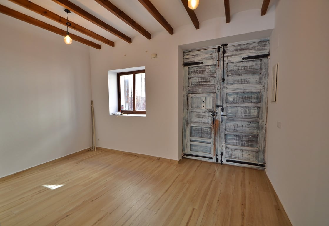 Entrada principal con puerta de madera rústica restaurada, vigas de madera a la vista y suelos de tarima clara en un espacio diáfano y luminoso.