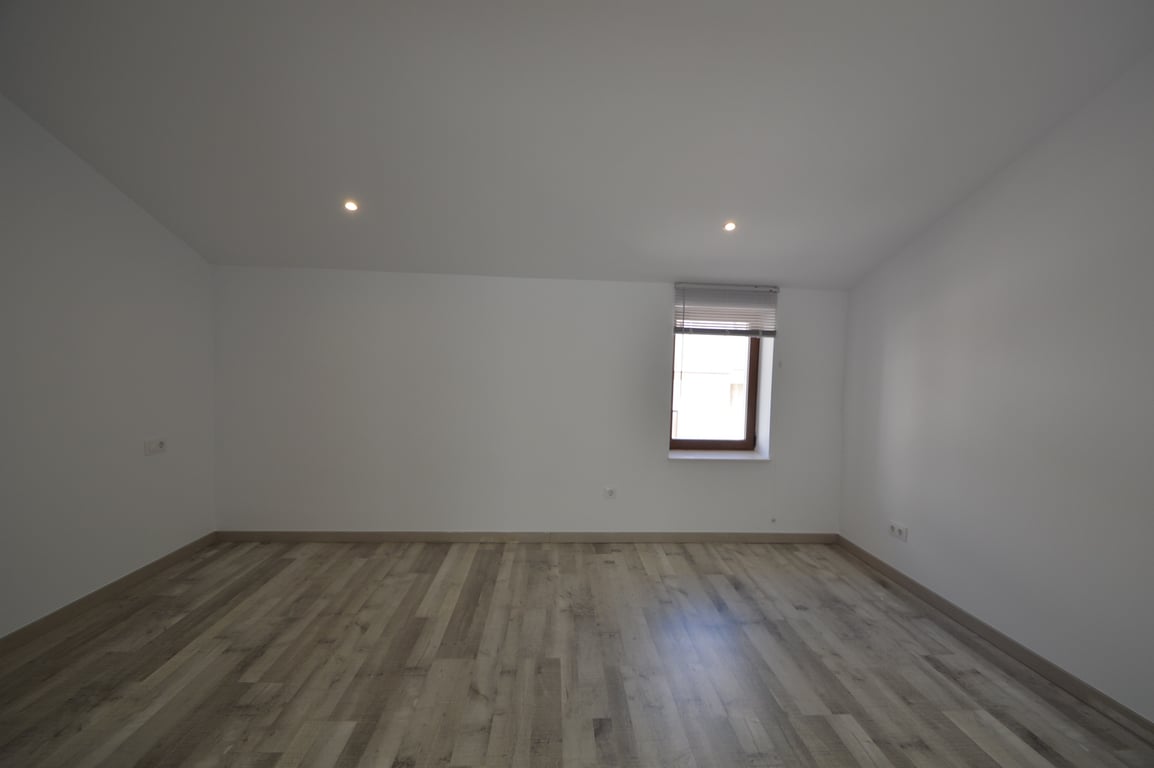 Habitación renovada con suelos de tarima clara, paredes blancas lisas e iluminación mediante focos empotrados y ventana con persiana.