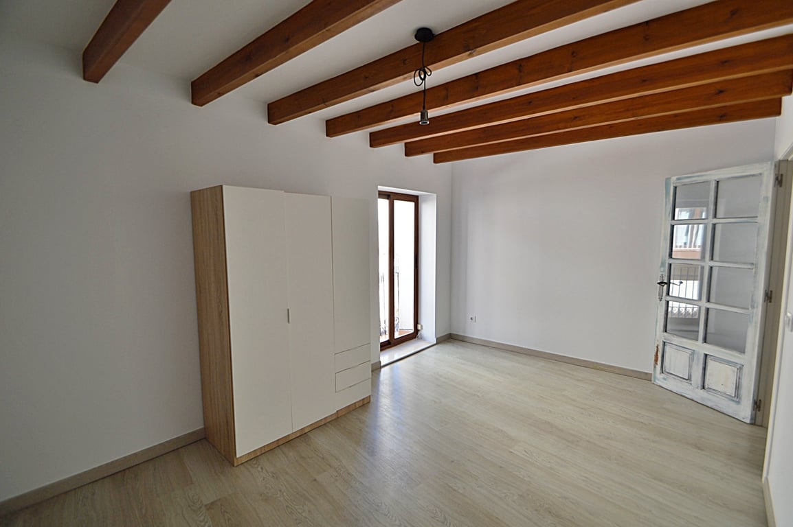Habitación renovada con vigas de madera originales, suelos de madera clara y acceso a balcón. Incluye un armario modular y una puerta artesanal de madera con cristales.