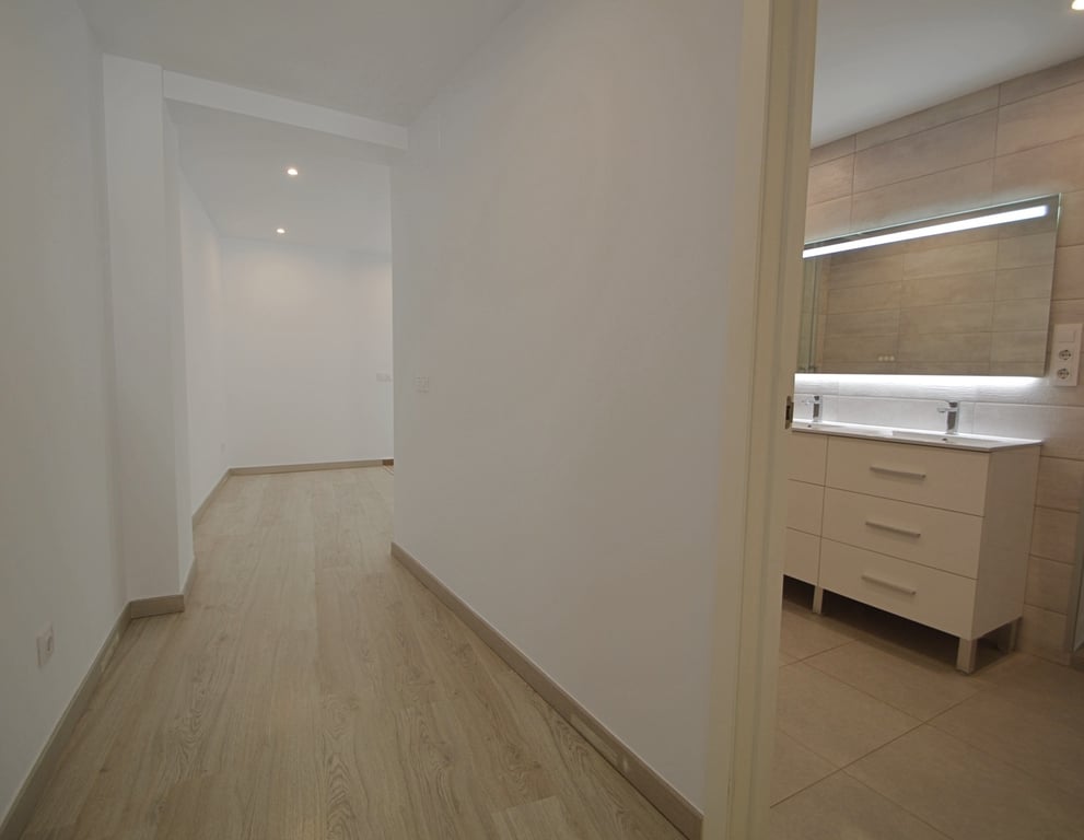 Pasillo con suelos de madera clara que conecta con un baño moderno equipado con lavabo doble, espejo retroiluminado y paredes alicatadas en tonos neutros.
